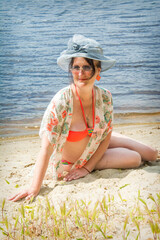 IIn summer, an imposing woman in a hat and glasses sits on the sand near the river.