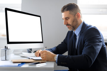 Serious middle aged businessman at table with computer with empty screen making notes, working in office, mockup