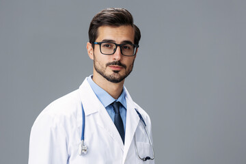 Fototapeta premium A serious doctor man in a white coat and eyeglasses and a stethoscope looks at the camera on a gray isolated background, copy space, space for text, health