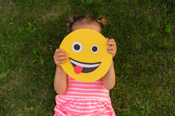 A close-up of crazy smile face making faces and showing his tongue in the hands of a small child lying on the grass of a lawn in a park