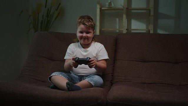 Boy enthusiastically plays game holding joystick in his hands while sitting on sofa in room in evening.