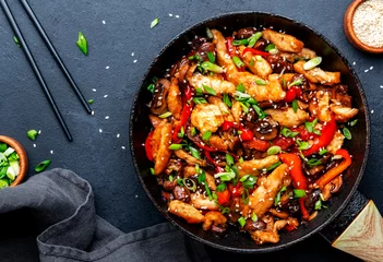 Foto auf Acrylglas Scharfe Chili-pfeffer Asian cuisine stir fried chicken, paprika, mushrooms, chives with sesame seeds in frying pan. Black kitchen table background, top view