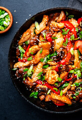 Asian cuisine stir fried chicken, paprika, mushrooms, chives with sesame seeds in frying pan. Black kitchen table background, top view