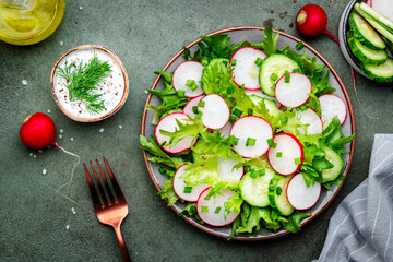 Healthy crispy salad with radishes, cucumbers, lettuce and chives with yogurt dressing, green stone table background, top view