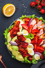 American Cobb salad with grilled chicken, cherry tomatoes, boiled eggs, bacon, avocado and lettuce, black stone table background, top view