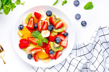 Fruit and berry salad with fresh strawberries, blueberries, banana, kiwi, orange and mint leaves, white table background, top view
