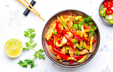 Asian cuisine stir fried chicken breast, red paprika and zucchini with sesame seeds, lime and soy sauce in ceramic bowl. White table background, top view