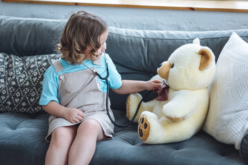 Cute little girl in a beautiful dress paying doctor at home with her teddy bear, listening to...