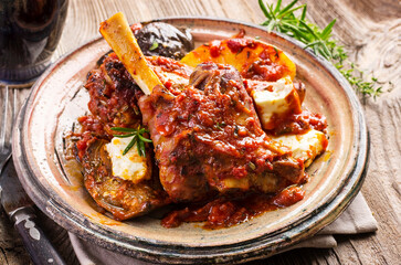 Traditional Greek braised lamb knuckle with feta cheese and eggplant in spicy tomato sauce served as close-up on a rustic design plate