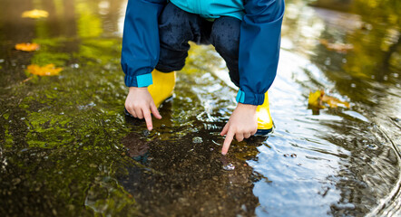 Adorable toddler boy wearing yellow rubber boots playing in a a puddle on sunny autumn day in city park. Child exploring nature. Fun autumn activities for kids.