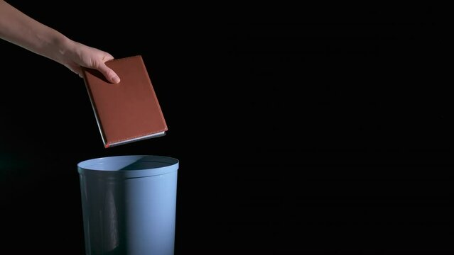 Bin with outdated book. A view of person hand throwing the book into the plastic bin on the black background.