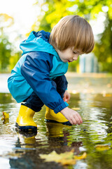 Adorable toddler boy wearing yellow rubber boots playing in a a puddle on sunny autumn day in city park. Child exploring nature. Fun autumn activities for kids.