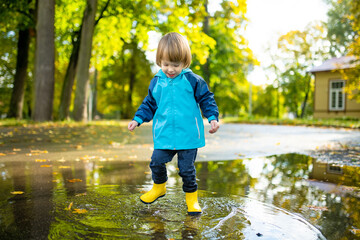 Adorable toddler boy wearing yellow rubber boots playing in a a puddle on sunny autumn day in city park. Child exploring nature. Fun autumn activities for kids.