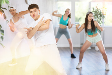 Fototapeta premium Positive smiling male teenager learns social dance, perform movements in choreographic class in company of peers. Young girls in ball cap and guy repeat movements, train in spacious training hall.