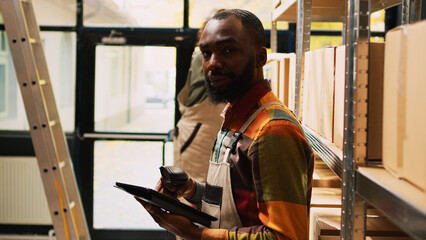 African american man scanning barcodes with tablet, working on quality control management in storehouse. Male employee checking stock of merchandise, using scanner in warehouse.