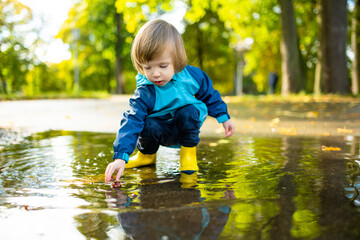 Adorable toddler boy wearing yellow rubber boots playing in a a puddle on sunny autumn day in city park. Child exploring nature. Fun autumn activities for kids.