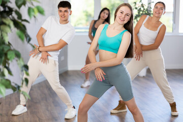 Fototapeta na wymiar Smiling teenage girl dancer practicing active vigorous dance with group of friends in modern studio..