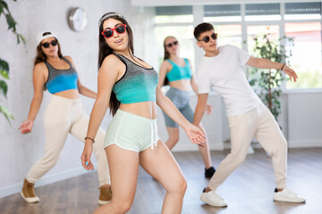 Fototapeta premium Female teen perform choreographic exercises and teach energetic mobile social dance shuffle with friends. Young girls in dark glasses and ball cap and guy repeat movements, train in spacious studio