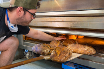 Male employee baker of bakery in workshop workshop of artisan bakery baking loaves in oven