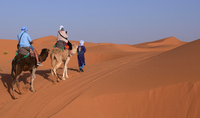 Tourists ride camels through the sand dunes in Sahara Desert. Berber man in national clothes...