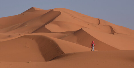 Fototapeta na wymiar Young female tourist going among deep orange - red dunes at sunrise in Sahara desert. Desert landscape with huge dunes. Travel in Morocco. Adventure vacation in desert.