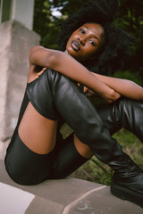 Fototapeta premium Fashion outdoor street style portrait Beautiful young African American woman posing outside on urban city landscape summer day wearing total black high leather boots.