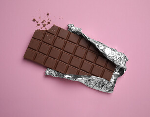 Fototapeta na wymiar Bitten milk chocolate bar wrapped in foil on pink background, top view
