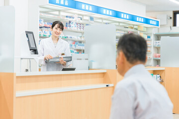 病院・薬局の待合室で問診票を書く高齢者男性と薬剤師
