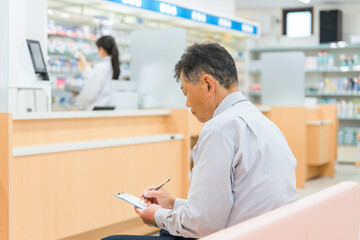 Fototapeta premium 病院・薬局の待合室で問診票を書く高齢者男性と薬剤師 