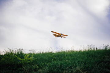 A yellow biplane flying over a populated area and a forest, spraying fertilizers for farm fields or chemicals against mosquitoes and other insects