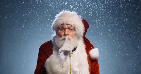 Old man in santa clause clothing looking at camera, winking and gesturing silence sign, isolated on snowy blue background - christmas spirit concept close up 