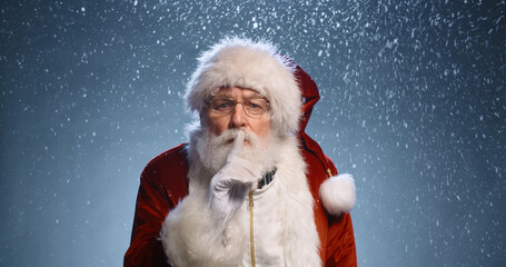 Old man in santa clause clothing looking at camera, winking and gesturing silence sign, isolated on...