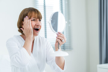 Caucasian senior woman looking at mirror after apply lotion on face. 
