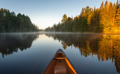The nose of a yellow canoe sticking out in front on a glass calm late with steam and sun light hitting the tips of trees  - obrazy, fototapety, plakaty