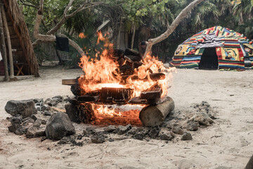 Bone fire burning with hot stones and logs in sandy tropical garden with indigenous hut in the...