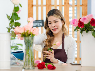 Asian happy cheerful professional female florist designer shop owner in apron sitting smiling using scissors cutting spikes decorating branch of red roses before selling in floral garden store studio