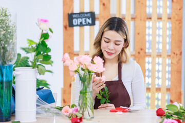 Asian happy cheerful professional female florist designer shop owner in apron sitting smiling using scissors cutting spikes decorating branch of red roses before selling in floral garden store studio