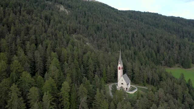 Aerial drone view of an old secluded mountain top church in the Italian alps surrounded by a forrest of trees 01