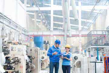 American male engineer and female manager Checking job details with listnote holding a radio Wear a helmet and uniform. in a large plastic and steel industry There are machines working around.