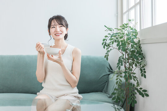 家のソファでヨーグルト・乳製品を食べるアジア人女性（ビフィズス菌・乳酸菌・乳製品・腸活）
