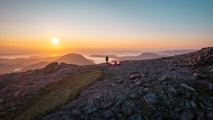 Fototapeta na wymiar Hiker enjoys sunset while camping on top of An Teallach, witnessing the beauty of Scottish highlands landscape.