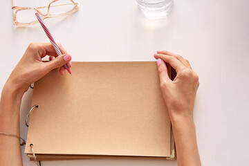 Woman is writing by her left hand on a craft paper paper. Mockup album. Glasess and glass of water. Conceptual photo of international left handers day. Copy space. Top view