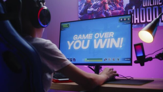 Happy gamer in headphones wins a round in online video game on home personal computer. Schoolboy plays third person shooter. Desk illuminated by RGB LED strip light. Gaming at home concept. Back view.