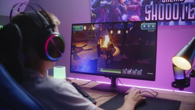 Young boy puts on headphones and starts play third person shooter. Gamer enjoys online video game on home personal computer. Desk illuminated by RGB LED strip light. Gaming at home concept. Back view.