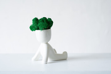White ceramic figurine of human with colorful stabilized moss fro head is sitting whit hands back