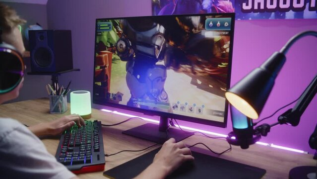Young gamer plays third person shooter and talks with teammates. Schoolboy in headphones enjoys online video game on home computer. Desk illuminated by RGB LED strip light. Gaming at home. Dolly shot.