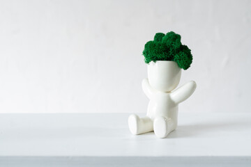 White ceramic figurine of human with colorful stabilized moss from head is sitting with hands up. 