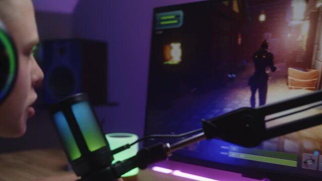 Gamer in headphones plays third person shooter on home computer and talks with teammates, uses streaming microphone. Schoolboy loses round in PvP multiplayer game. Esports remote tournament. Close up.