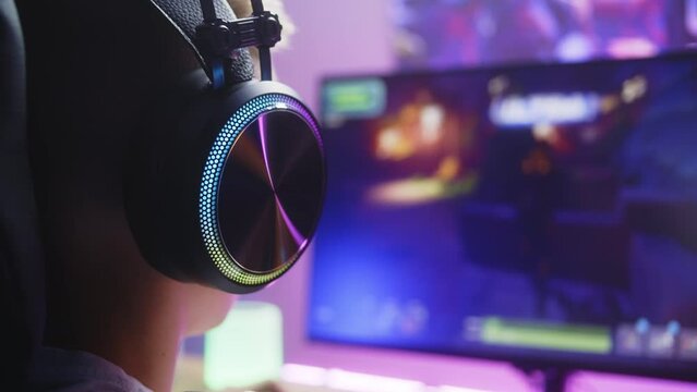 Young boy in headphones plays third person shooter. Gamer enjoys online video game on home personal computer. Desk illuminated by RGB LED strip light. Concept of gaming at home. Back view. Close up.