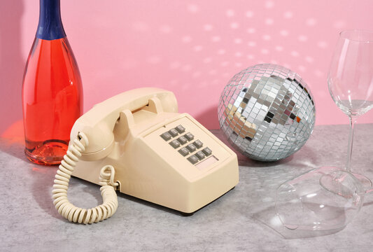A retro phone with gray buttons stands among a bottle of champagne, a glittering disco ball and glasses.